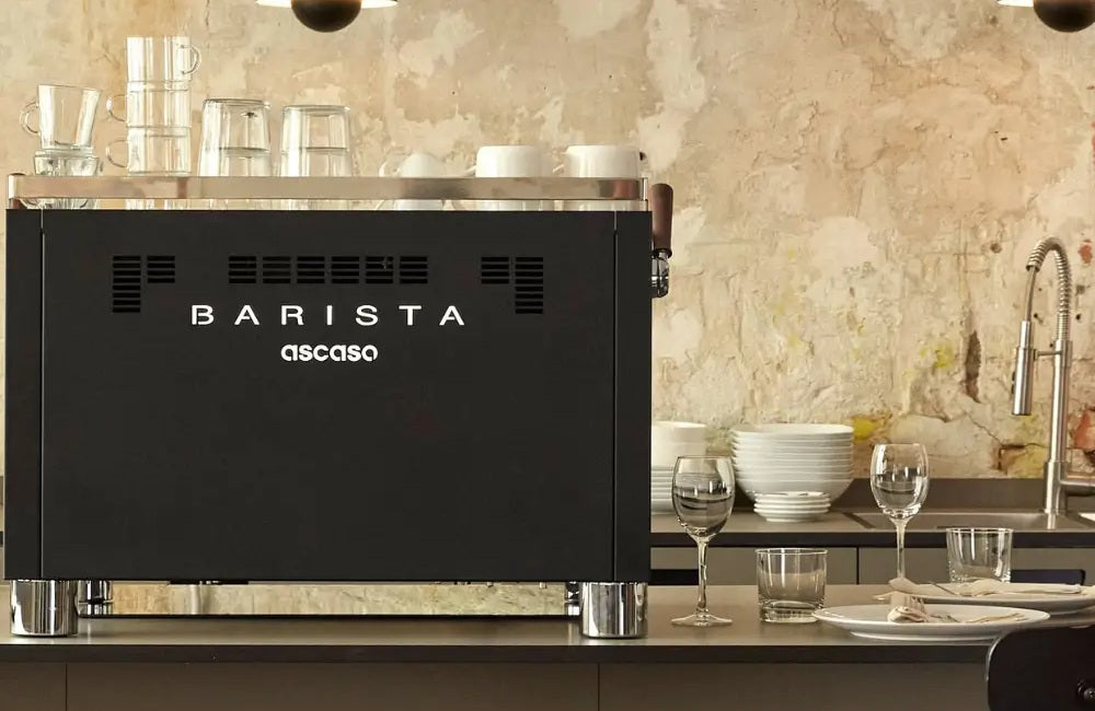 Rear view of a black Ascaso Barista T commercial espresso machine on a counter in a stylish, industrial-chic cafe setting.