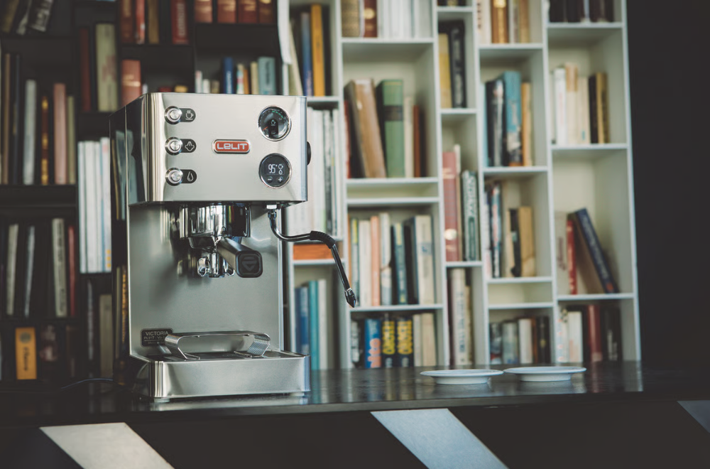 Lelit Victoria espresso machine with digital PID display turned on, sitting on a black table in a home library setting with bookshelves in the background.