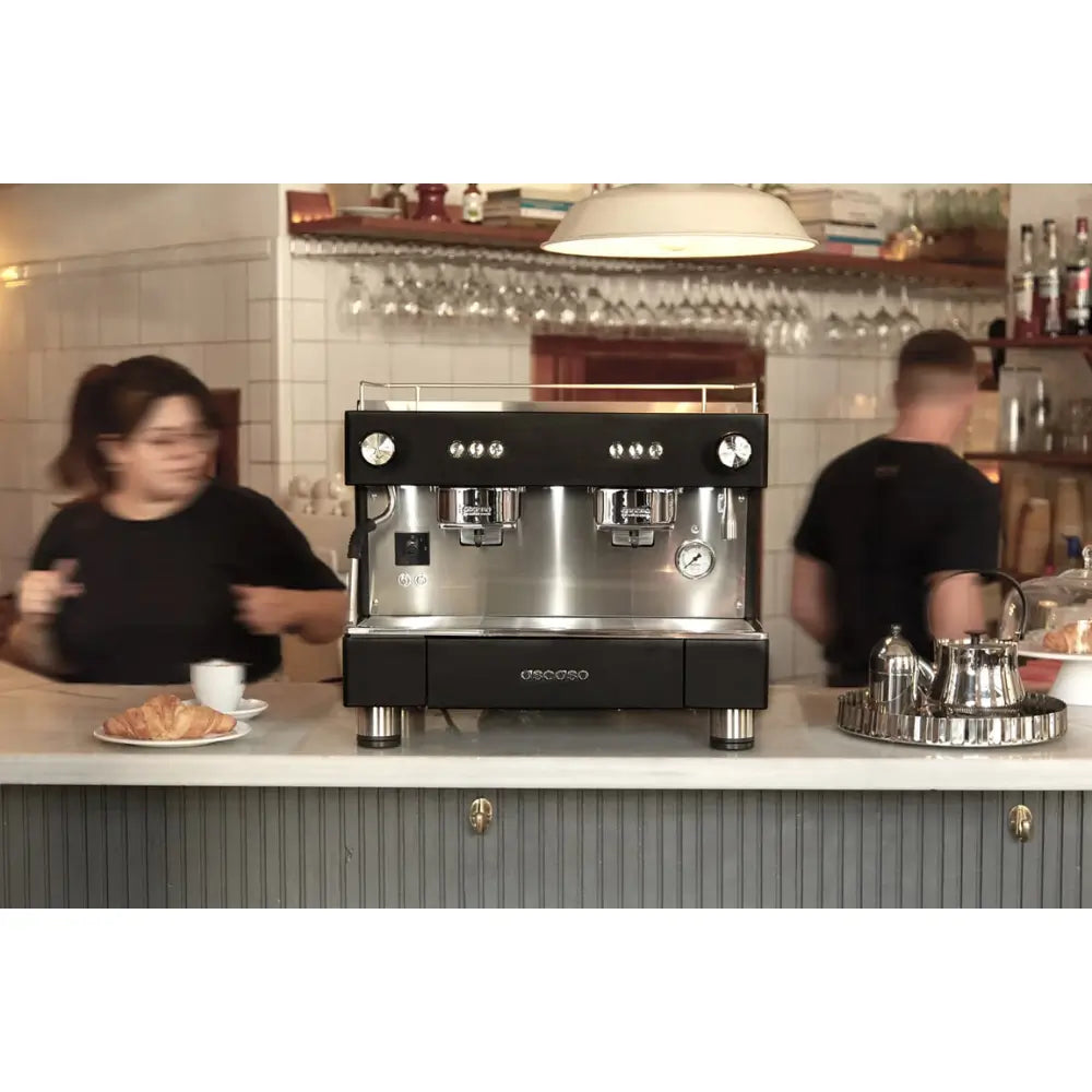 Front view of the Ascaso BAR 2 Group espresso machine in a café, with baristas in the background and a cup of coffee served on the counter.