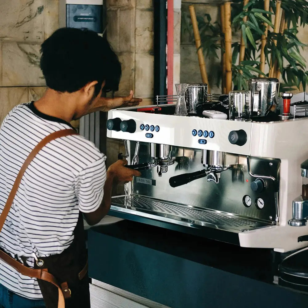 Barista in a striped shirt and leather apron pulling an espresso shot on the white Iberital Lanna Intenz machine in a cafe.