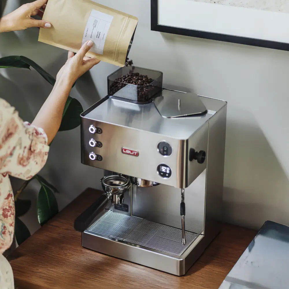 Person pouring fresh coffee beans into the hopper of a Lelit Kate espresso machine in a home kitchen.