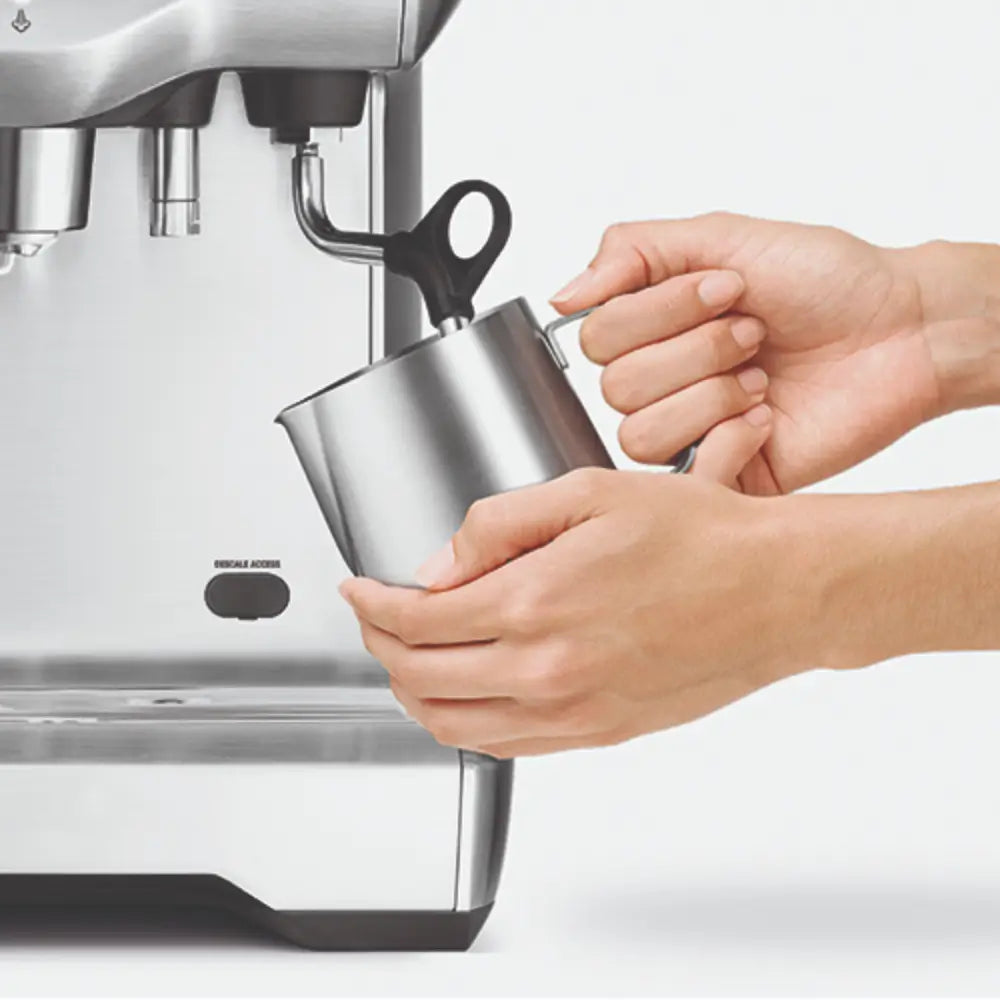 Hands using the powerful steam wand on the Sage Dual Boiler to texture milk in a stainless steel pitcher for microfoam and latte art.