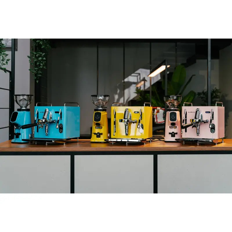 Lineup of several Sanremo Cube R espresso machines in bright colours (yellow, blue, pink) on a professional counter.