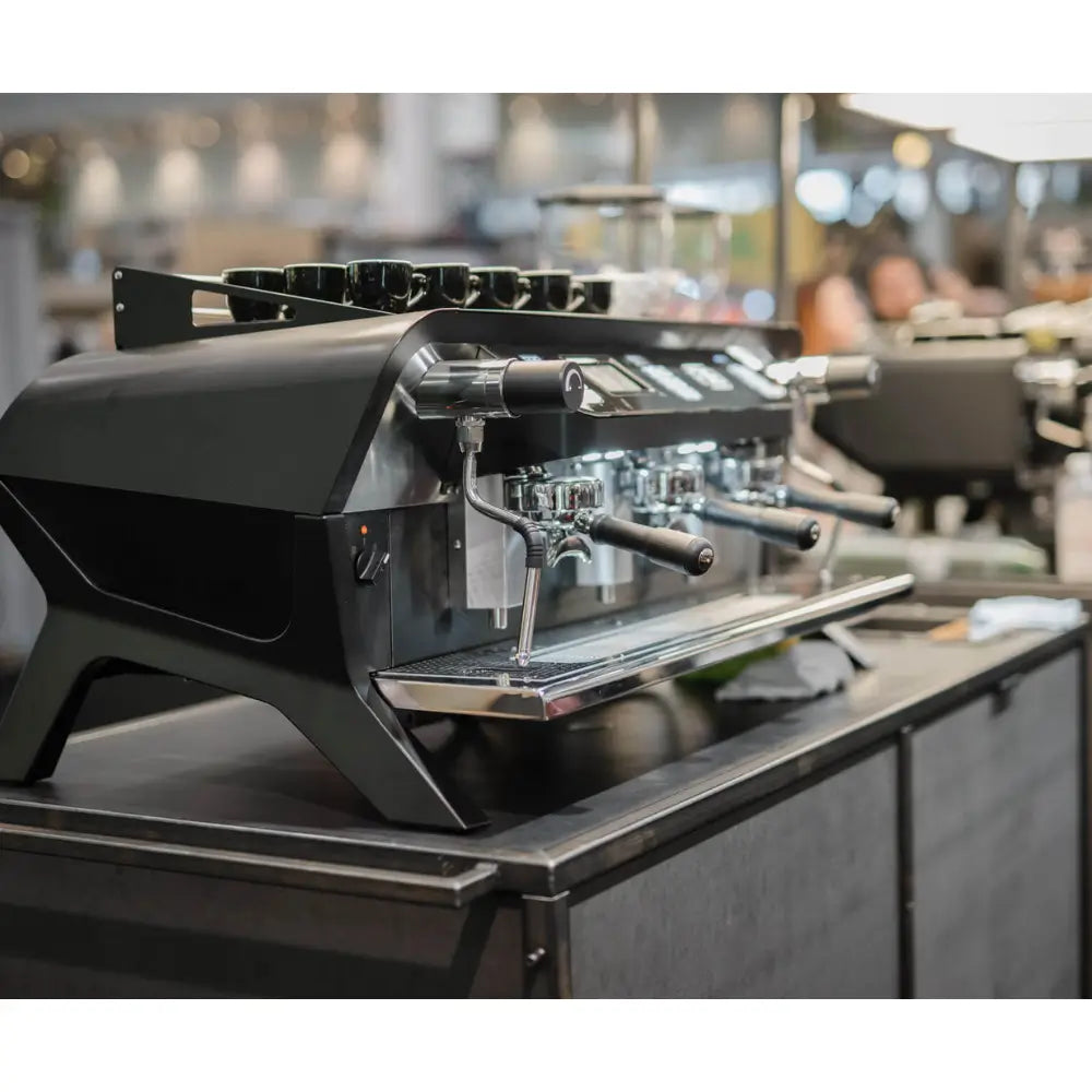The sleek black Sanremo F18 Multi Boiler on a café bar with cups warming, featuring the Sanremo signage in the background.