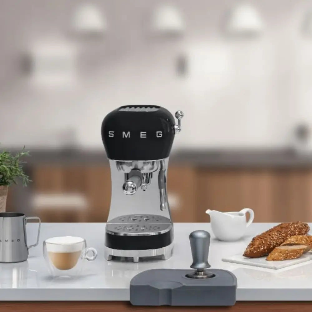 SMEG ECF02 espresso machine in black on a kitchen counter with a tamper station and fresh bread.