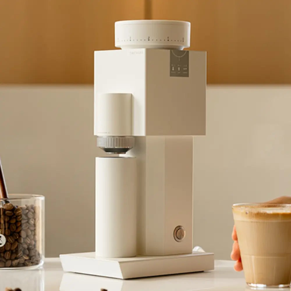 A white Timemore Bricks 01S electric coffee grinder on a kitchen counter next to a glass of iced latte, showcasing its clean, square design suitable for modern home coffee bars.