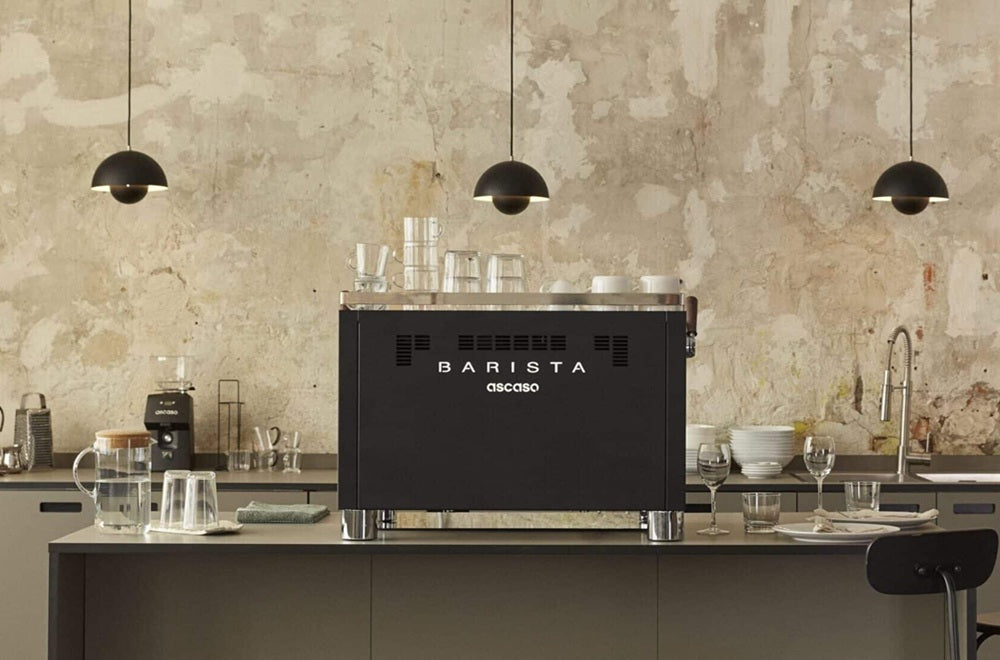 Rear view of a black Ascaso Barista T commercial espresso machine on a counter in a stylish, industrial-chic cafe setting with pendant lighting.