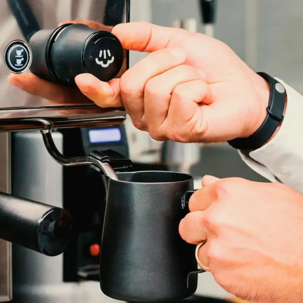 Close-up of a barista's hands using the steam wand and a black milk pitcher on a professional espresso machine.