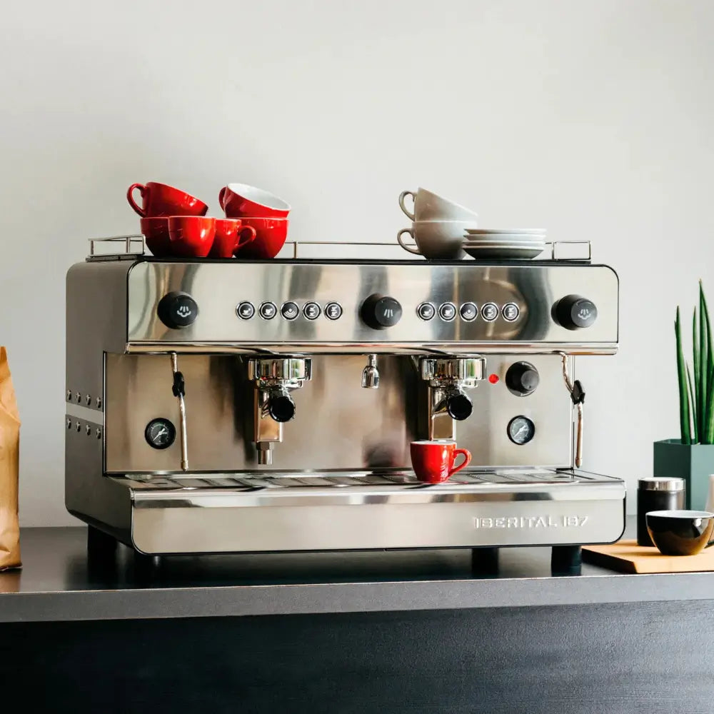 Iberital IB7 2 Group stainless steel espresso machine on a black counter with red and gray coffee cups stacked on top.