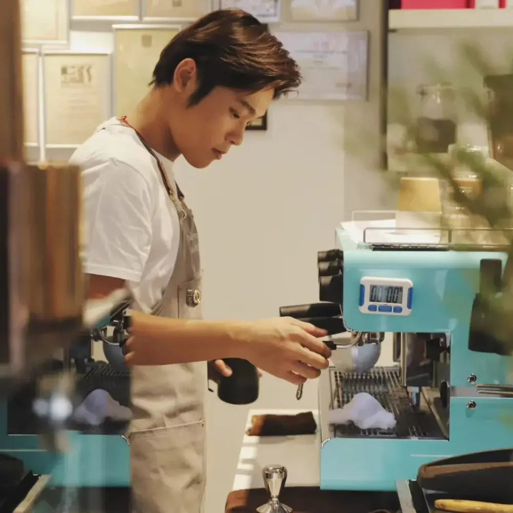 A professional barista in an apron operating a portafilter on a light blue commercial espresso machine with a digital timer.