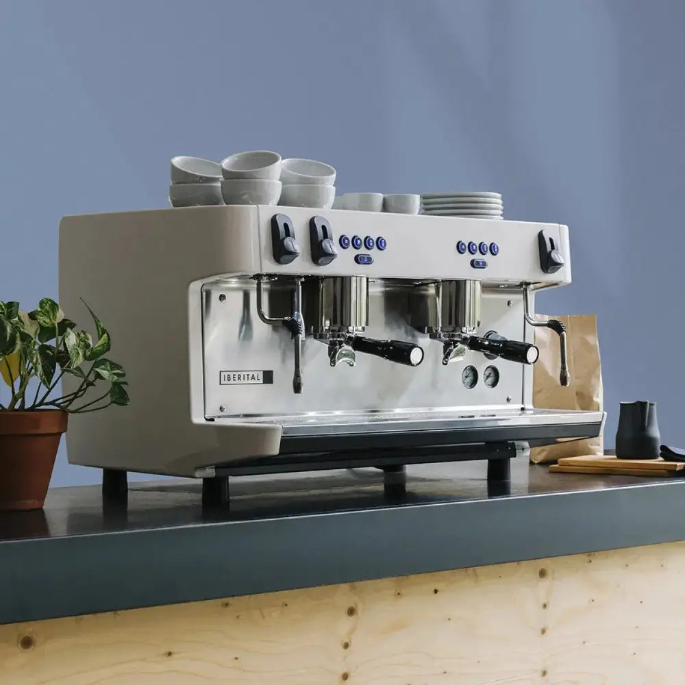 White Iberital Lanna Intenz espresso machine on a wooden and gray counter, styled with stacked cups and a potted plant.