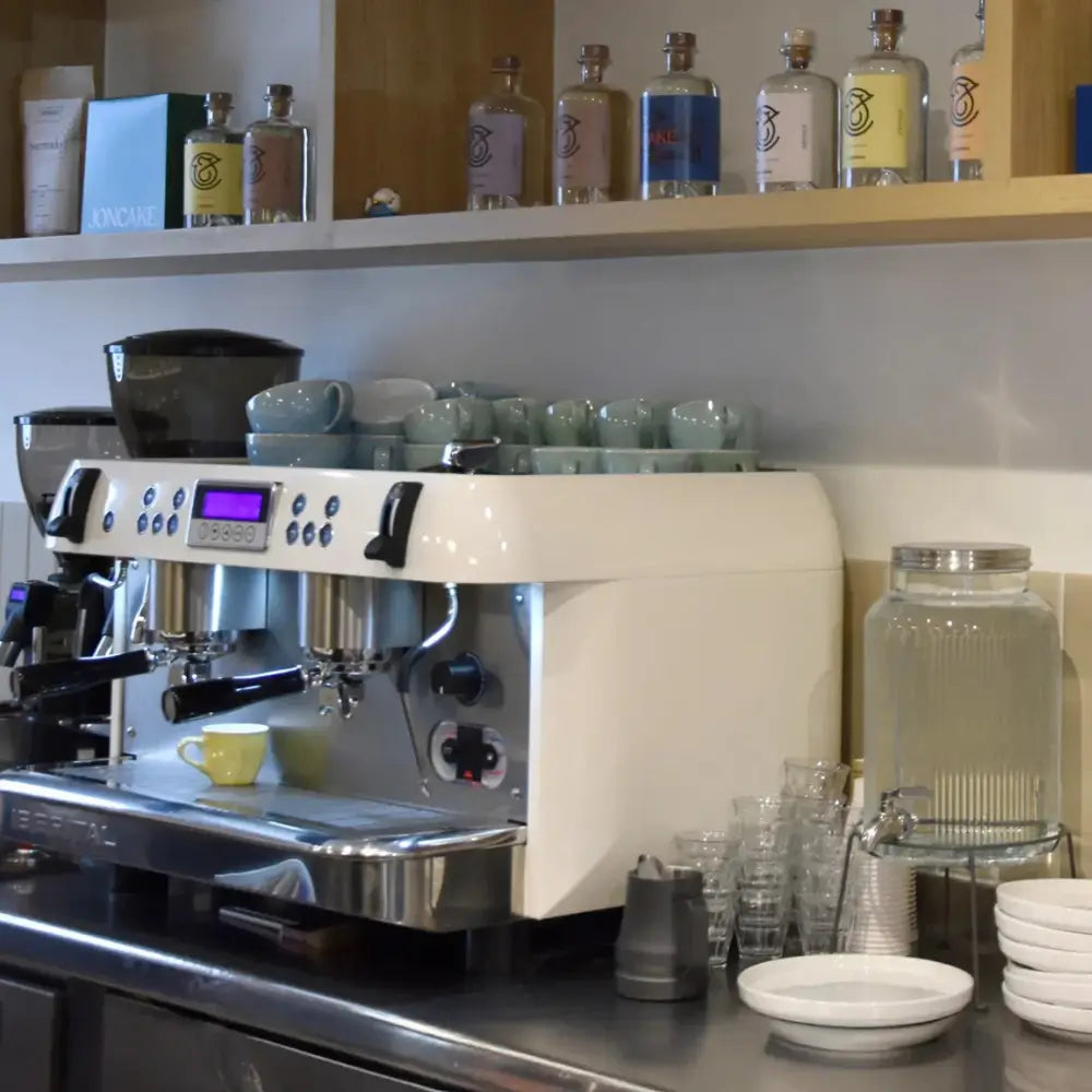 Wide shot of a cafe counter with the white Iberital New PID machine, surrounded by stacked cups, bottles, and a water dispenser.
