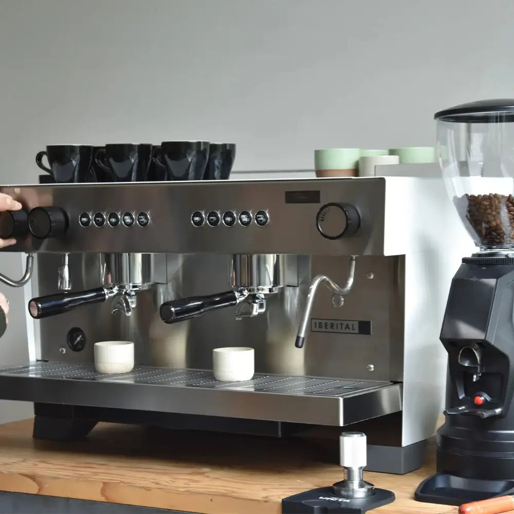 Iberital Referent 2 Group espresso machine alongside a coffee grinder, with cups stacked, on a wooden counter.