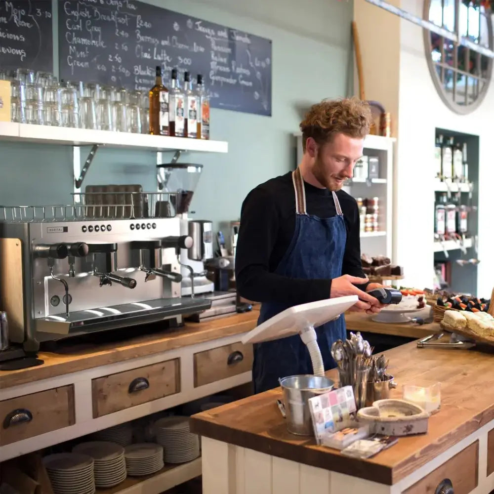Barista smiling while operating a point-of-sale (POS) terminal next to the large Iberital Tandem espresso machine in a bright cafe setting.