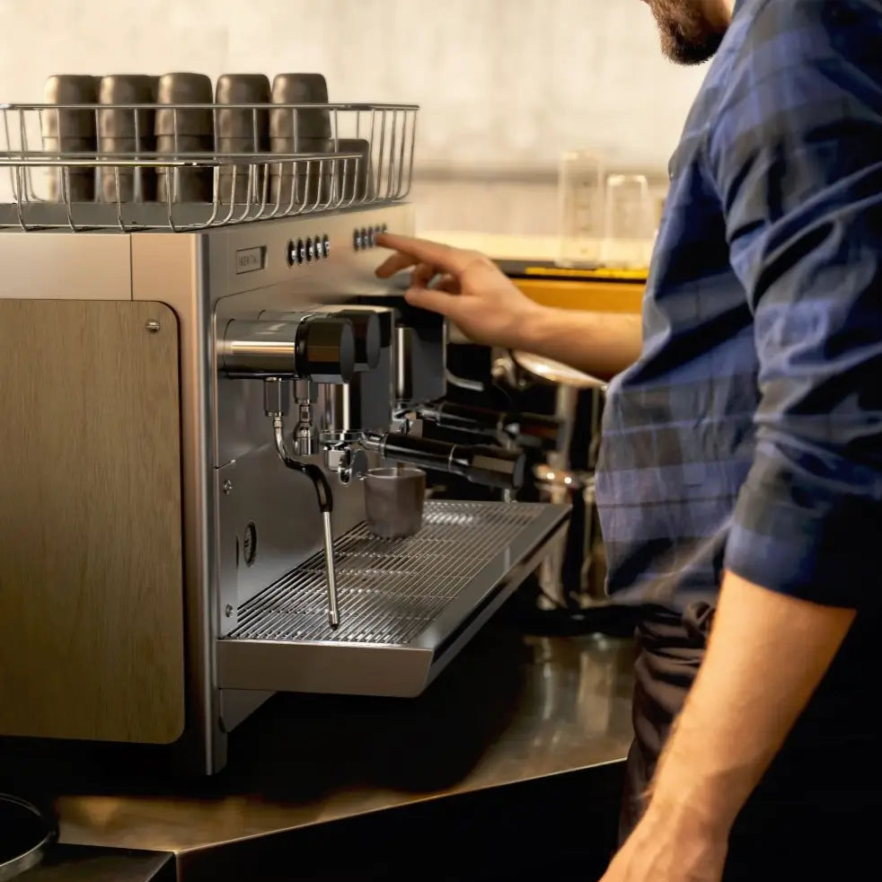 A barista in a blue plaid shirt presses buttons on the Iberital Tandem control panel to start a shot.