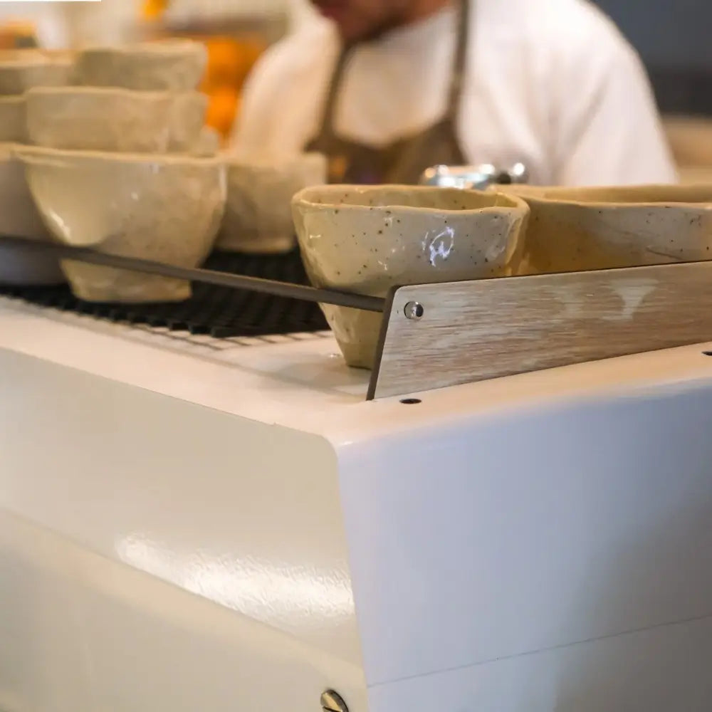 Close-up of handmade ceramic cups resting on the white Iberital Vista cup warming tray, with a barista blurred in the background.