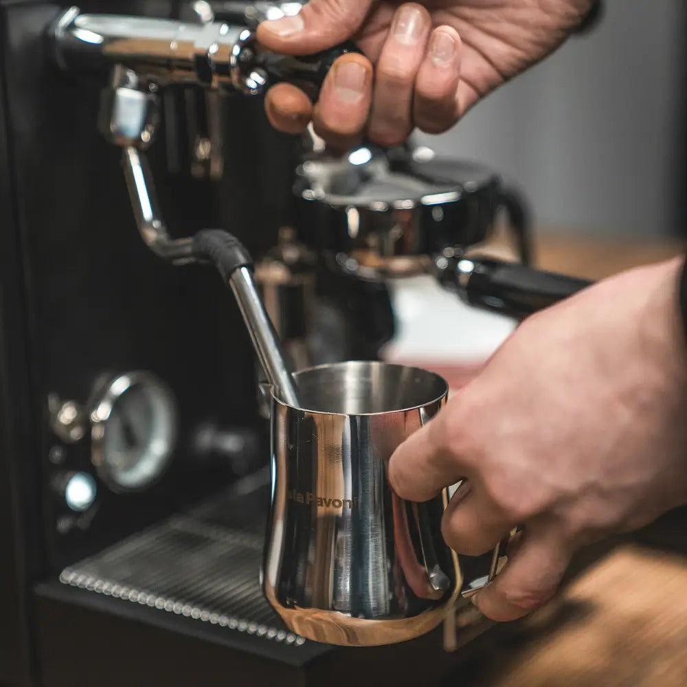 A hand using the steam wand to froth milk in a stainless steel jug on the La Pavoni Cellini Evolution.