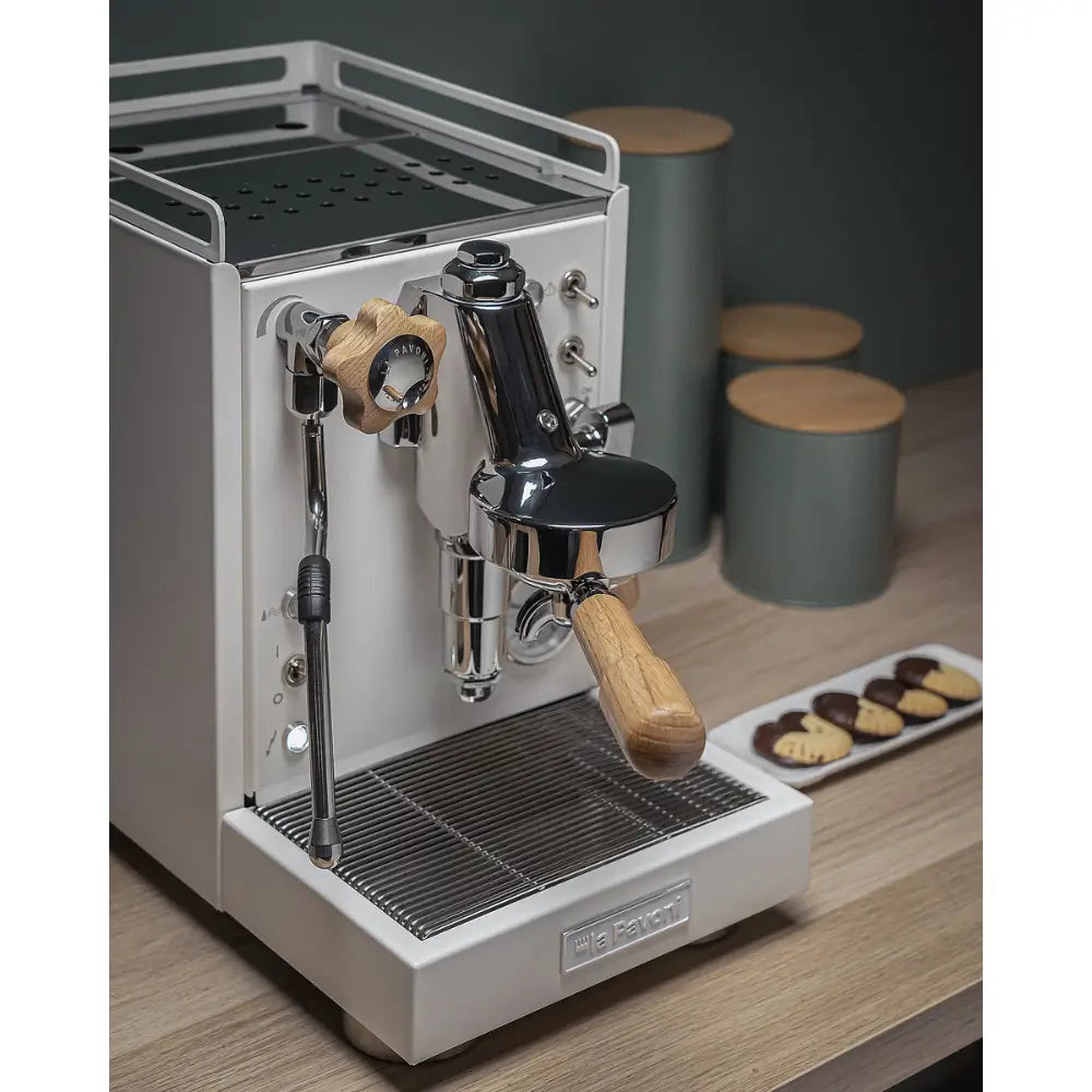 The La Pavoni Mini Cellini espresso machine on a wooden counter with a plate of cookies and coffee containers.