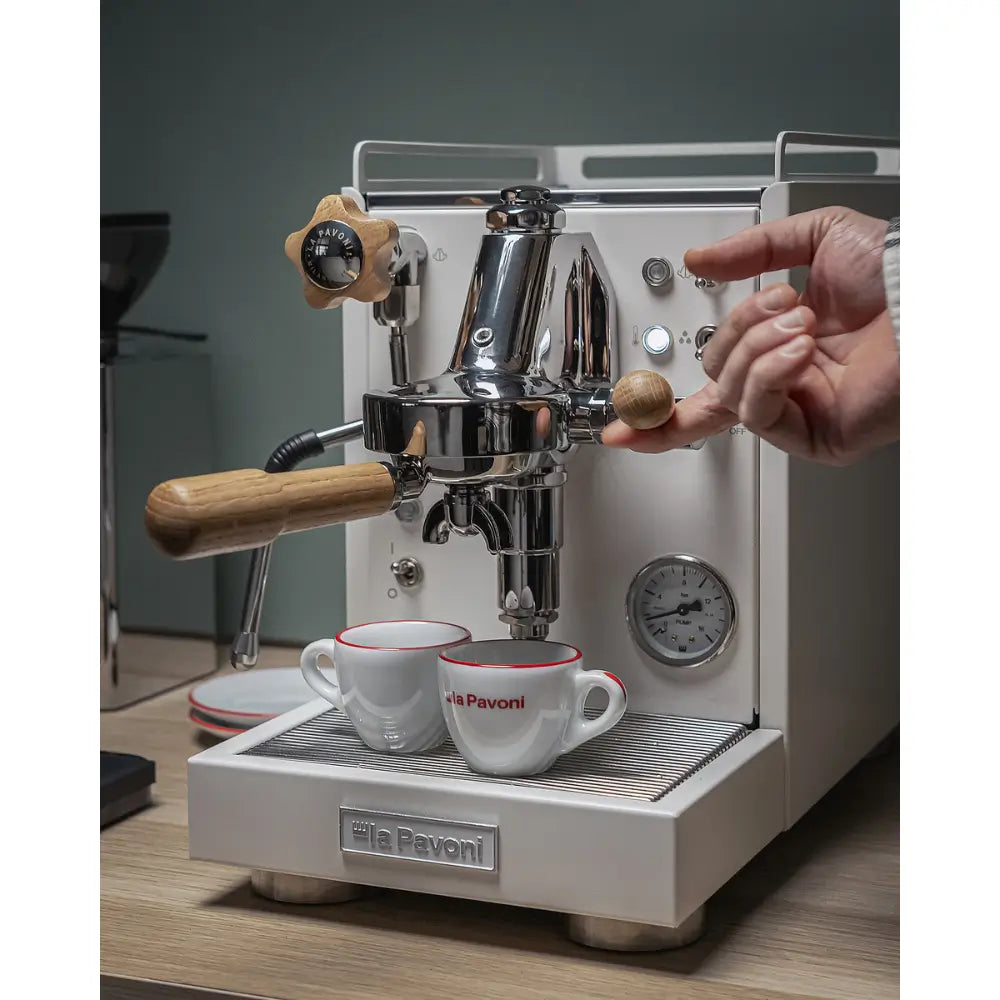 A hand operating a switch on the La Pavoni Mini Cellini, with espresso cups ready on the drip tray.
