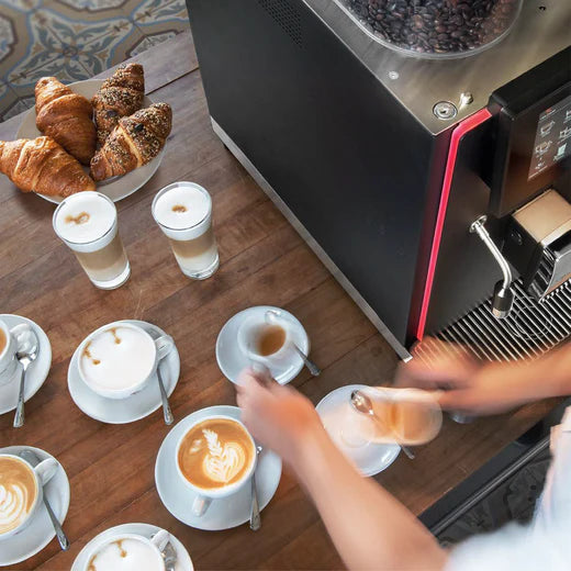 Barista preparing multiple espresso drinks beside the Rex-Royal S1 bean-to-cup coffee machine with pastries and lattes on a wooden counter