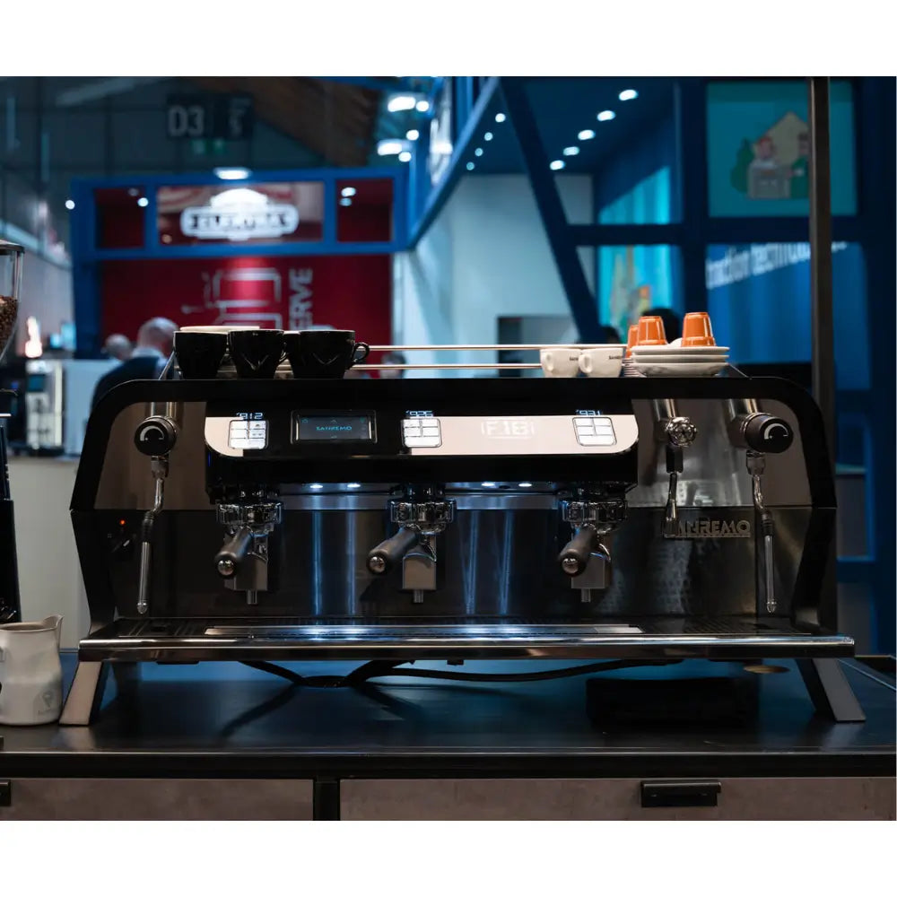 Front view of the black Sanremo F18 Multi Boiler showing the three group heads and chrome drip tray in a working cafe environment.