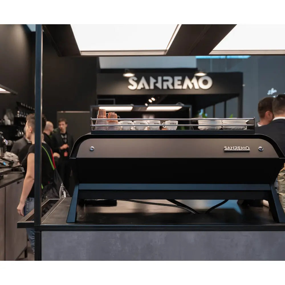 The sleek black Sanremo F18 Multi Boiler on a café bar with cups warming, featuring the Sanremo signage in the background.