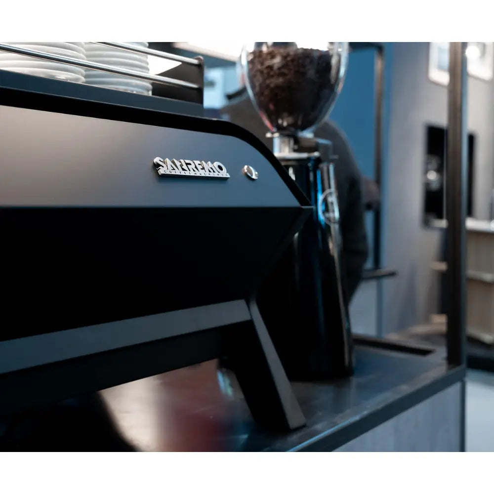 Close-up of the Sanremo logo on the F18 MB's rear panel, with a commercial grinder visible next to it.