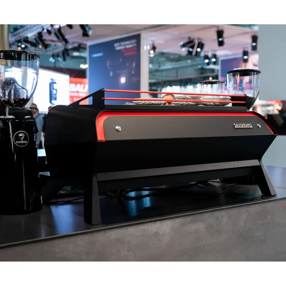 Lifestyle shot of the black Sanremo F18 Multi Boiler with red accent trim, sitting next to a grinder on a café counter.