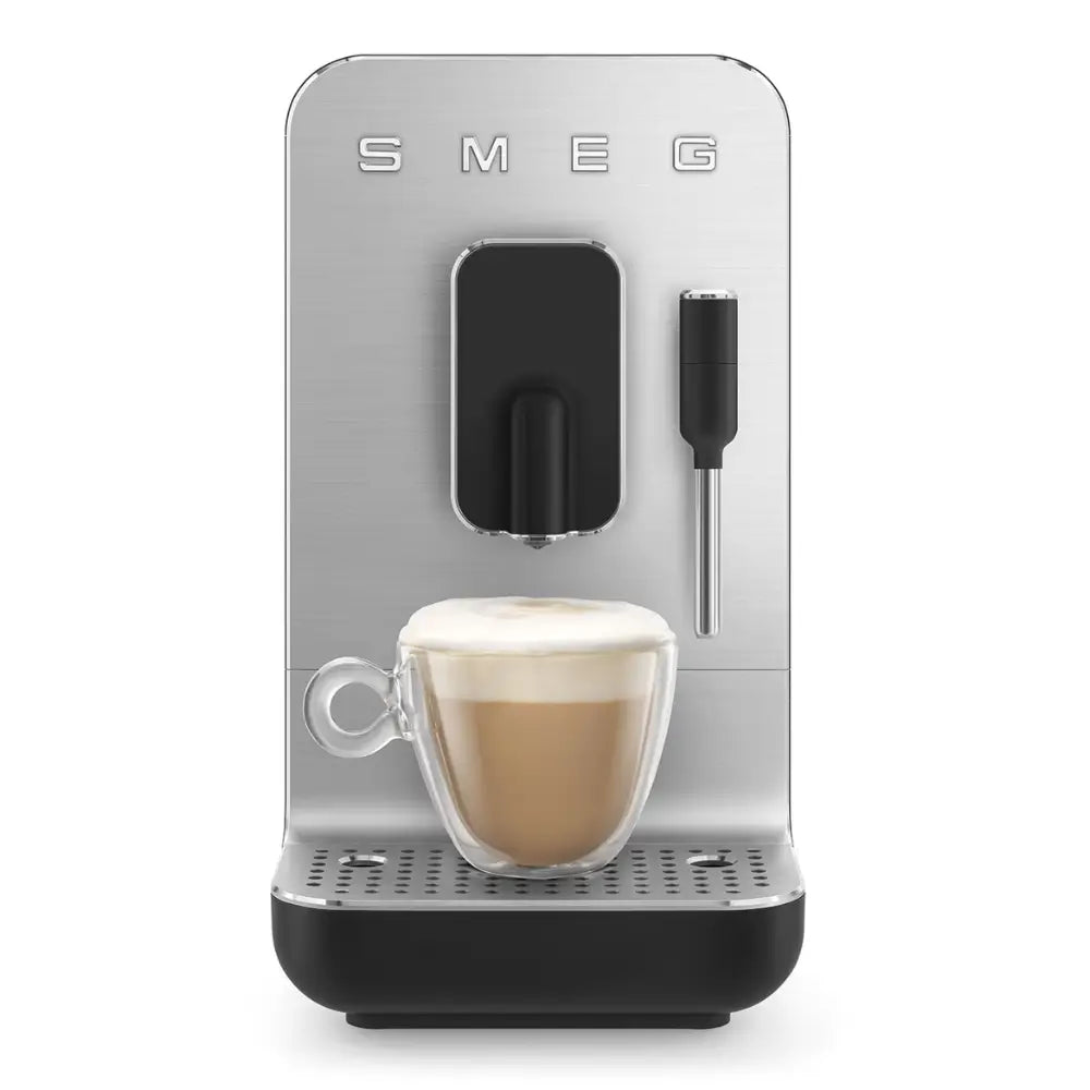 Front view of the SMEG BCC12 brewing a frothy cappuccino into a glass mug, using its steam wand.