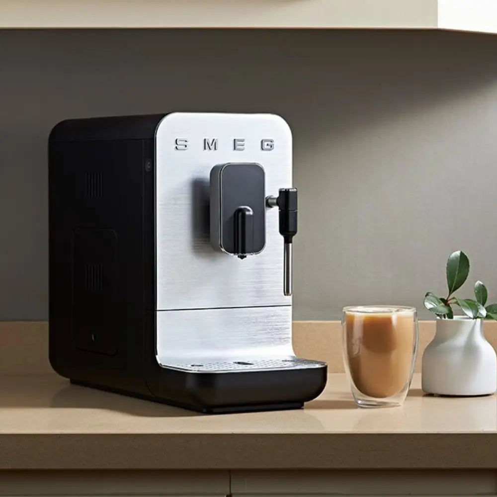 SMEG BCC12 bean-to-cup machine on a home countertop next to a fresh latte, showing its compact profile.
