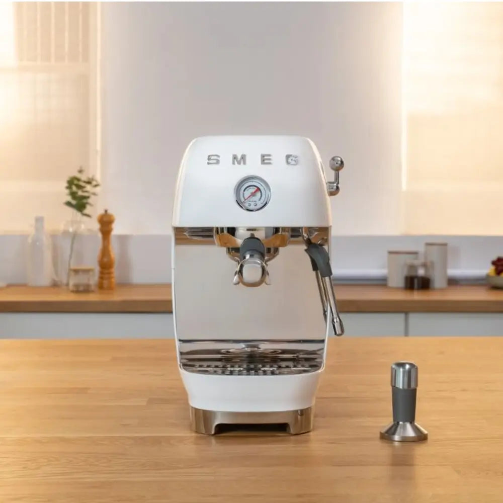 White SMEG ECF03 espresso machine sitting on a wooden kitchen countertop in a bright room, with a tamper beside it.