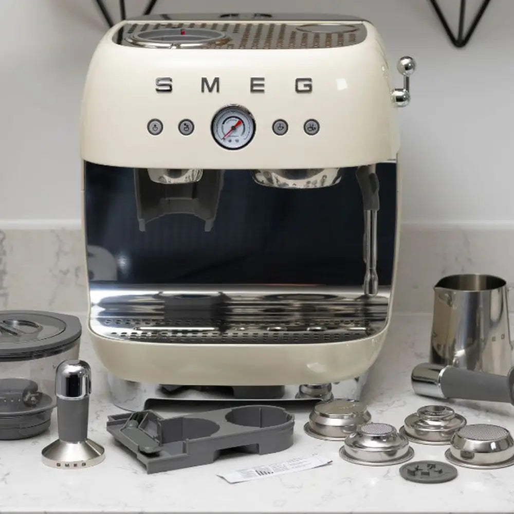 SMEG EGF03 in cream finish on a marble counter surrounded by its included accessories, such as the tamper and filter baskets.