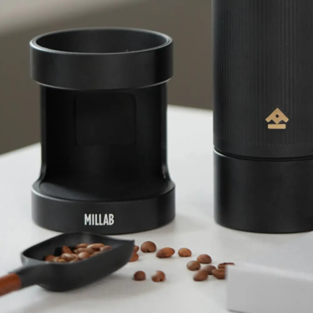 Close-up of the black Timemore x Millab grinder base and dosing cup next to a scoop of roasted coffee beans, showcasing the compact setup for home espresso or pour-over preparation.