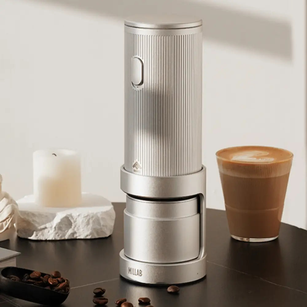 A silver Timemore x Millab wireless grinder placed on a table next to a glass of latte and decorative candles, illustrating a modern home cafe aesthetic.