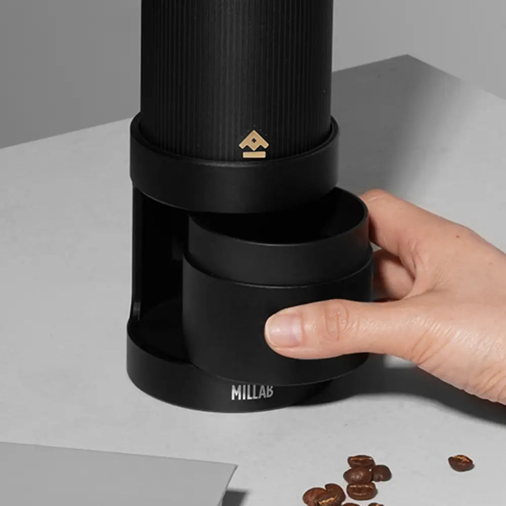 A user sliding the magnetic catch cup out of the black Timemore x Millab electric grinder stand, demonstrating the easy-access design for transferring ground coffee beans.